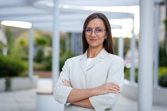 Businesswoman in glasses stands arms crossed, smiling slightly at camera. Office professional wears white blazer in bright workspace, conveying poise and approachable authority, with hands folded