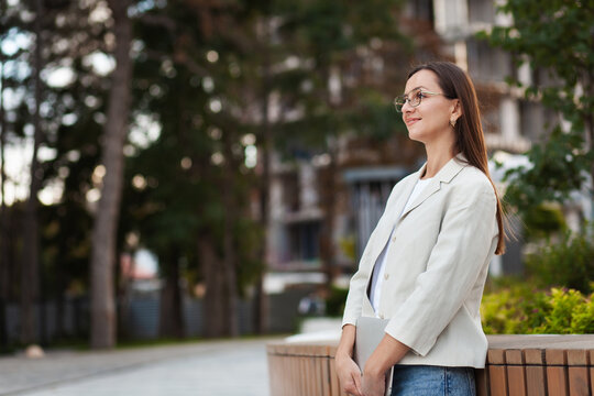 Professional woman smiles while standing on city sidewalk in white blazer and glasses. Urban career portrait features poised businesswoman beside landscaped greenery and contemporary architecture.