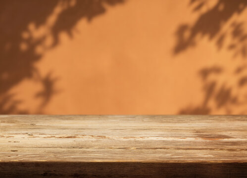 Bare wooden table fills foreground against orange backdrop patterned by blurred foliage shadows. Rustic tabletop provides inviting product display surface and ample space for merchandise.