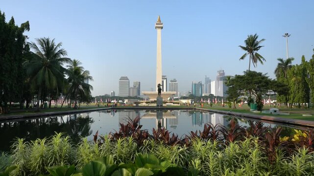 Jakarta, Indonesia - October 21, 2026: The National Monument stands above a landscaped park with a reflecting pool, tropical vegetation, and high-rise buildings in the background.