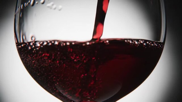 Slow motion close up of red wine pouring into stemmed glass with swirling liquid and bubbles on white studio background, glossy deep burgundy tones, intimate evening mood