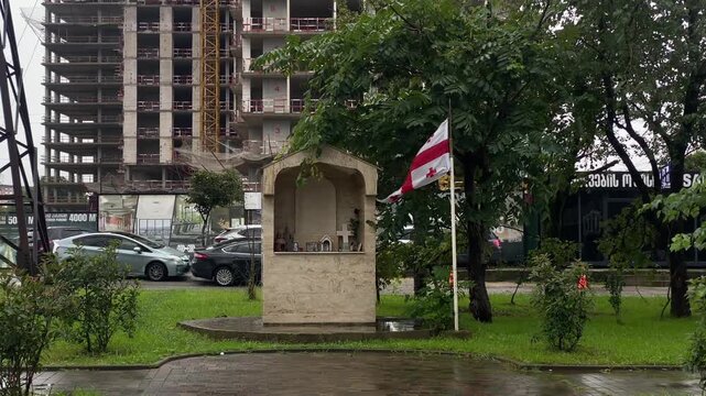 A small stone roadside shrine with a Georgian flag stands among greenery near parked cars and a high-rise construction site. Footage for cultural travel