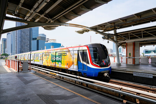 BTS Skytrain stopped at an elevated Bangkok station, with platform structures, city towers and advertising panels in a busy urban setting, Thailand, Bangkok, April 26, 2026.