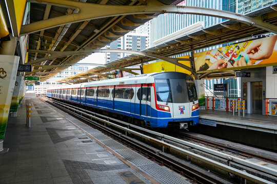 BTS Skytrain arrives at an elevated Bangkok station beneath a large geometric metal canopy, with platform signs and modern buildings surrounding, Thailand, Bangkok, April 26, 2026.