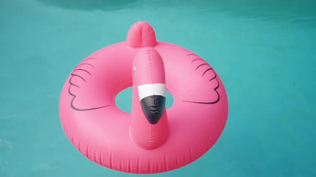 A pink inflatable flamingo pool float drifts slowly on calm turquoise water. Top view of the ring with painted wings and black beak. Water drops on vinyl. Bright summer vacation and pool party fun.