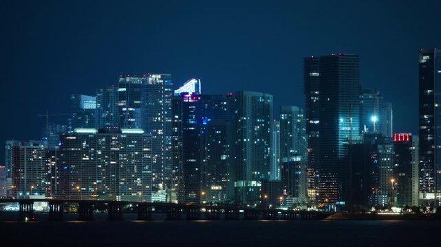 Night Miami downtown skyline with lit glass towers, red rooftop beacons and a construction crane above a long causeway bridge on dark bay water. Palm trees and street lamps line the shore.