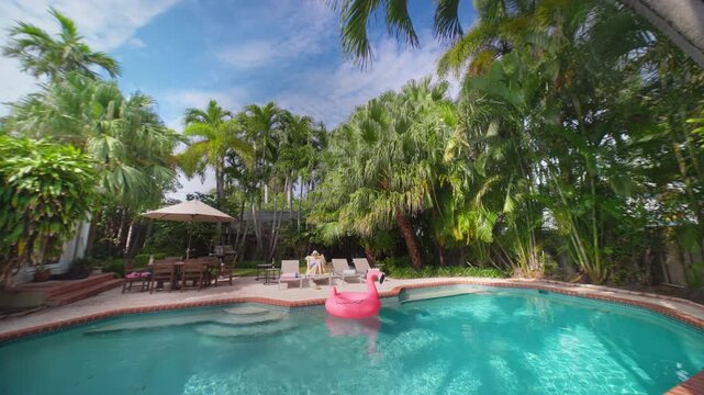 Beautiful girl relaxing by the pool on a sun lounger in a tropical backyard. Pink flamingo float on turquoise water. Tall palm trees, patio umbrella and dining table on a sunny summer day.