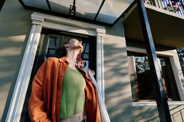 Fototapeta premium Young woman in an orange shirt and green top stands by a house door, face lifted to warm sunlight. Low angle view. Concept of relaxed summer lifestyle at home.