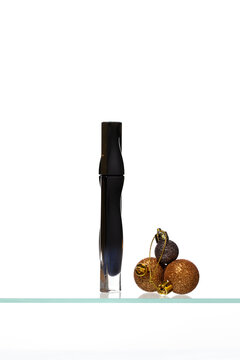 Sleek black mascara tube standing vertically next to bronze and gold glitter Christmas ornaments on a transparent glass shelf against a clean white background.