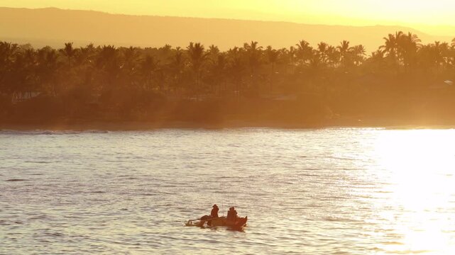 Small fishing boat with three anglers drifts on shimmering ocean at golden sunset off the palm lined coast of Big Island Hawaii with mountain ridges and tropical shoreline glowing in warm light. 