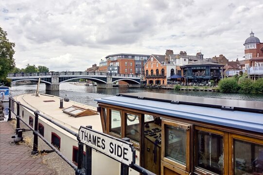 Windsor Bridge and traditional narrowboat moored along the scenic River Thames in Windsor, Berkshire, England, featuring historic architecture and a cloudy sky.