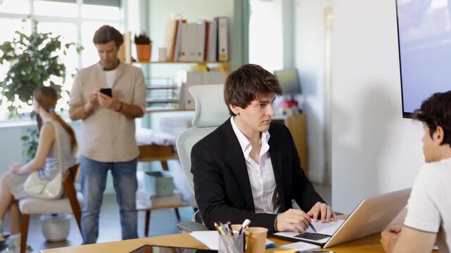 Adult male bank employee sits at desk with laptop and discussing with young male customer 