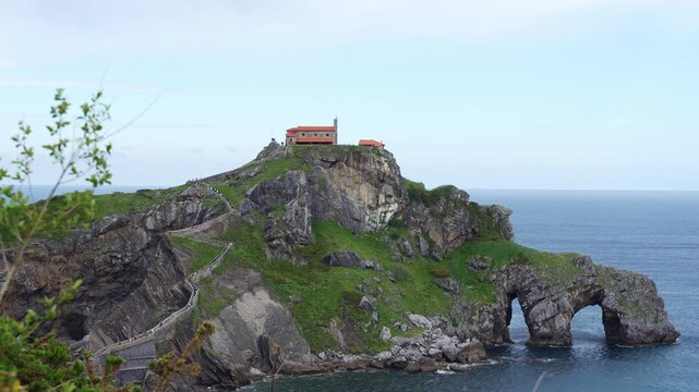 Rocky islet of San Juan de Gaztelugatxe with green slopes, natural stone arches, a long stone stairway and the hermitage on top surrounded by blue sea in Bizkaia, Basque Country, Spain