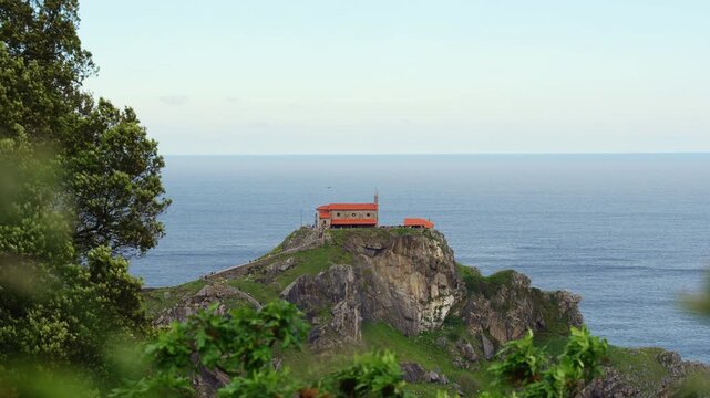 Telephoto view of the red roofed hermitage on top of the rocky Gaztelugatxe islet, framed by green trees and blurred leaves with the calm open sea behind in the Basque Country, Spain