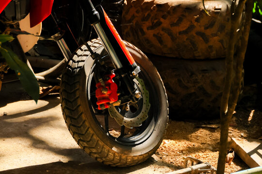 Red motorcycle wheel with knobby tire, disc brake, and suspension beside outdoor work area