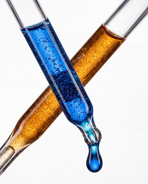Two crossed glass pipettes filled with blue oxygen serum and golden oil liquid with microbubbles, dripping a blue drop isolated on a white background.