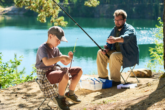 Caucasian father teaching his child boy fishing near lake shore, preparing tackle outdoors. Concept suitable for family bonding, outdoor recreation and hobby gear advertising