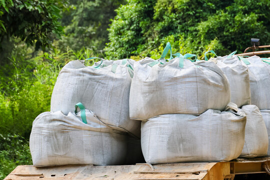 Stacked woven sacks filled with agricultural material rest on wooden truck bed beside lush tropical vegetation, showing rural transport and manual loading work
