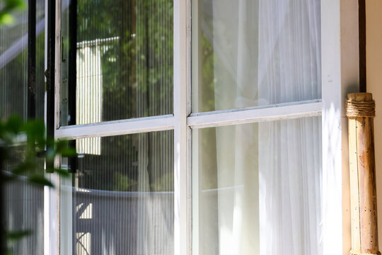 White window with sheer curtain, wooden column, and leafy reflection in bright residential sunlight