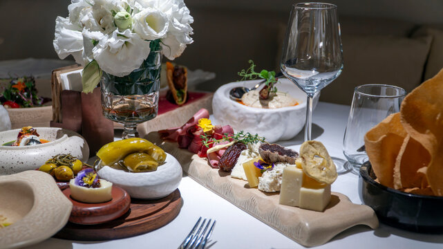 Charcuterie board with cheese cubes, cured meats, dates and edible flowers, with olives, white flowers and wine glasses on a restaurant table in Turkey