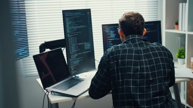 Young Caucasian male programmer writes code using multiple screens in home office workspace. AI development, cybersecurity, network database system, software engineering, remote tech startup concept.