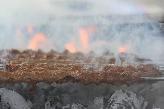 Traditional Adana kebab skewers cooking over open fire flames and charcoal grill with smoke. close-up view of minced lamb meat barbecue