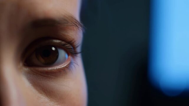 Brown eye shifts gaze and briefly lowers upper eyelid in extreme close-up, with detailed eyelashes, textured skin and cool blue reflections against softly blurred dark background