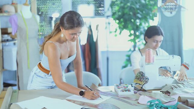 Focused young seamstress stitching linen fabric on sewing machine while female colleague drawing sketches by hand on paper sheet at worktable, creating bespoke garment together in workshop 