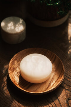 A round white natural soap sits on a wooden dish in warm sunlight