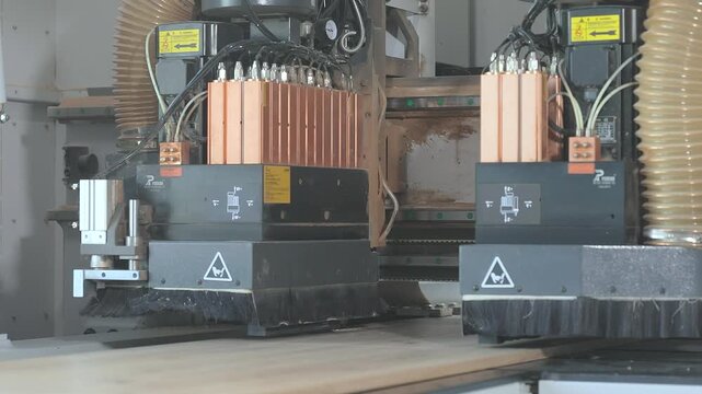 An automated CNC milling and drilling machine processes wooden panels at a furniture factory. A close-up of a dual-head CNC milling machine with dust extraction hoses processing particleboard panels