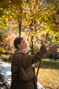 Woman takes selfie with smartphone beneath golden autumn trees in sunlit park. Digital lifestyle, social media, self expression, urban nature highlight creativity, leisure, seasonal wellbeing