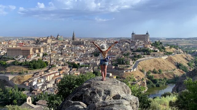 happy female tourist with open arms standing on rock overlooking toledo medieval city skyline and alcazar in castile la mancha spain