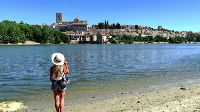 female tourist with straw hat standing on riverbank looking at zamora cathedral and medieval city skyline in castile and leon spain