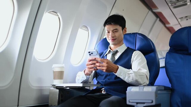 A young Asian executive smiles while using his smartphone in business class. Modern corporate travel, mobile connectivity, and remote work lifestyle during a flight.