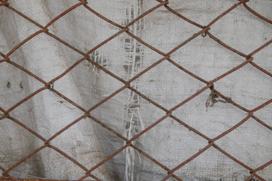 Rusty metal wire chain link fence texture close up with white polypropylene fabric background. Industrial security mesh net pattern backdrop for construction, safety, barrier, and vintage designs.