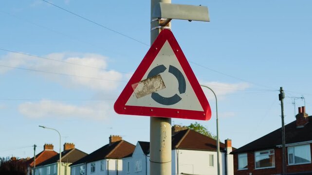 Roundabout ahead warning road sign on residential street. Weathered triangular traffic sign with red border and damaged symbol on metal pole. Suburban houses and streetlights beneath blue sky with