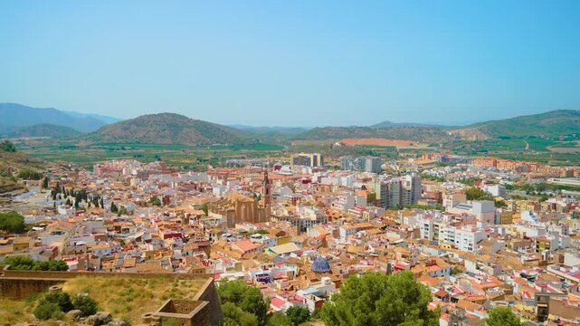 Wide Spanish cityscape with colorful rooftops and historic church under clear blue sky. Green hills and farmland beyond urban district. Mediterranean architecture in sunny Spain