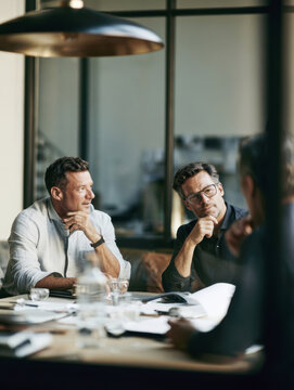 Two focused middle aged businessmen sitting around a modern conference table engaged in deep professional discussion during a strategic corporate meeting in a stylish glass walled office room