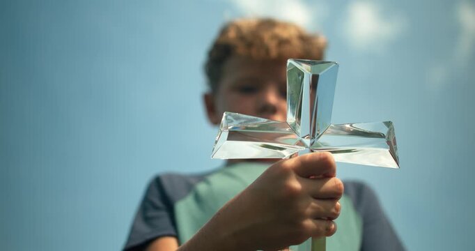 Low angle view of a young boy holding a clear glass cross toward the camera beneath a blue sky.