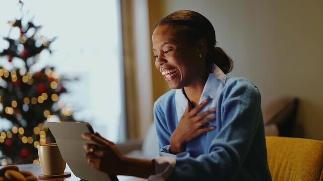 Smiling young woman holding a tablet and eating a cookie at the dining table, enjoying digital entertainment during a cozy Christmas break at home