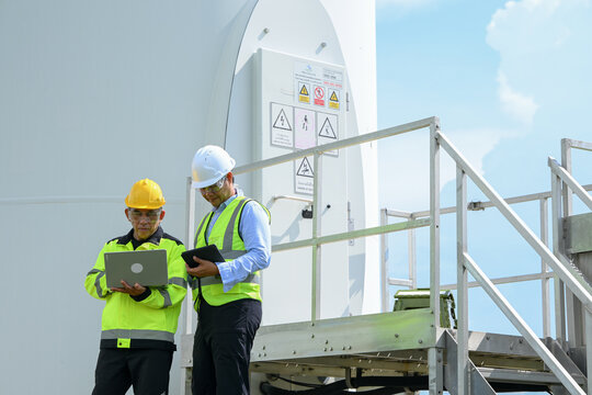 Electrical engineer and worker inspect industrial equipment beside wind turbine tower, reviewing safety information outdoors under bright blue sky with focused expressions