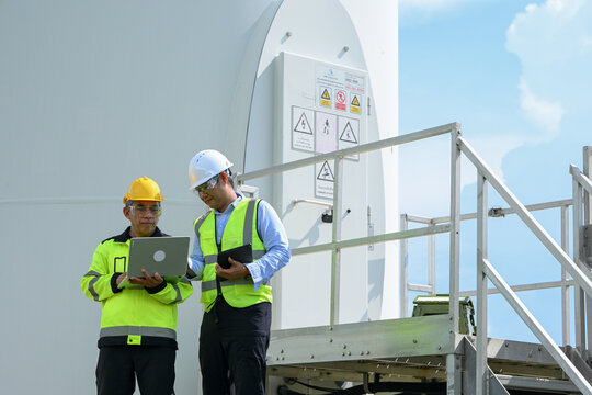And keywordsElectrical engineer inspecting wind turbine equipment with tablet beside industrial tower outdoors, wearing protective helmet and reflective vest during routine maintenance work bright
