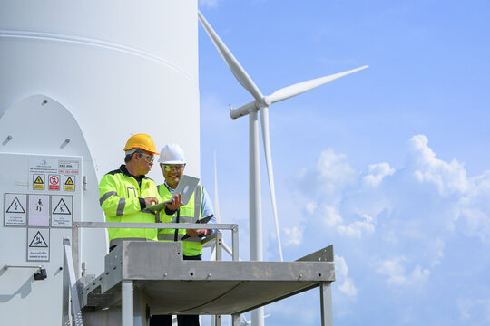 Wind turbine electrical engineer technician industrial worker safety helmet safety vest digital tablet renewable energy wind power facility maintenance platform turbine tower blue sky cloud clean
