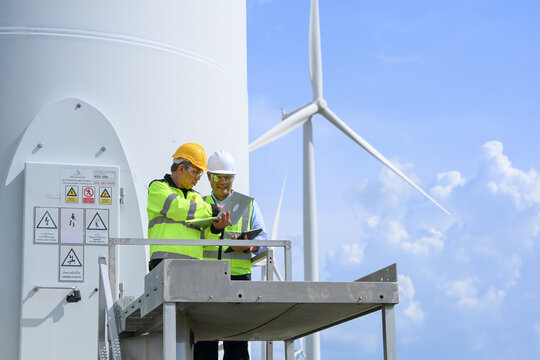 Electrical engineer and technician inspect wind turbine equipment on elevated platform, promoting renewable energy maintenance and workplace safety under bright blue sky