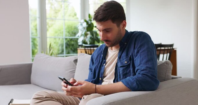 Man typing on a smartphone on a sofa in a bright living room
