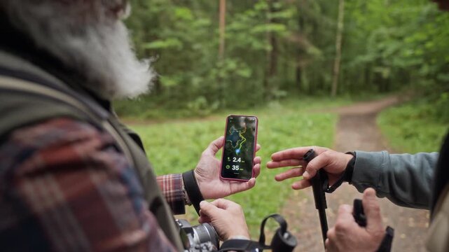 Two senior Caucasian men checking route on smartphone navigation app while hiking in forest. Useful for active retirement, travel tech, and outdoor lifestyle ads