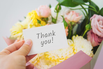 Female hand holding white card with handwritten text Thank you near pastel bouquet of pink roses, yellow chrysanthemums in pink wrapping, concept of gratitude, appreciation, gift, flower delivery © Evgeniia Primavera
