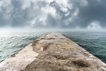 Naklejka premium A old concrete pier extends into a calm sea under a dramatic storm sky.