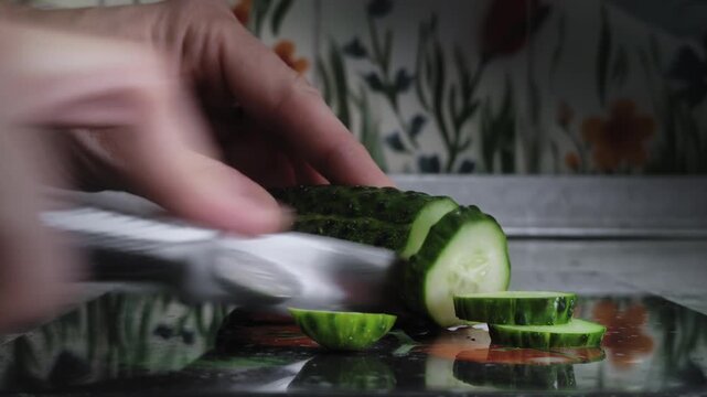 Male hand using metal knife slices cucumber, which lies on glass cutting board. Healthy food preparation for meal ingredient.