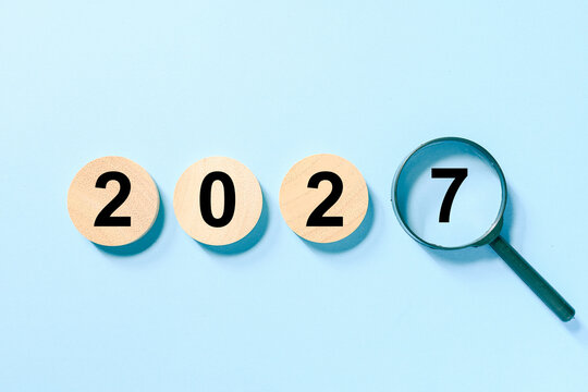 Wooden circles showing 202 and a magnifying glass highlighting 7 create a 2027 new year concept on a blue background, symbolizing planning, forecasting, research, and new opportunities.